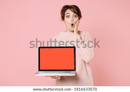 Shocked young woman 20s in knitted casual sweater hold laptop pc computer with blank empty screen mock up copy space, put hand on cheek isolated on pastel pink background. People lifestyle concept.