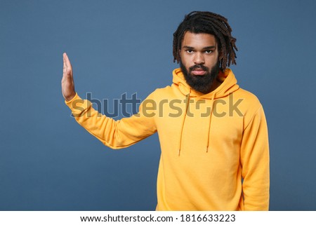 Serious young african american man with dreadlocks in casual yellow streetwear hoodie isolated showing stop gesture with palm aside on blue background studio portrait. People emotion lifestyle concept