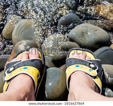 Women's feet in beach flip-flops on large sea pebbles near the water on the shore.