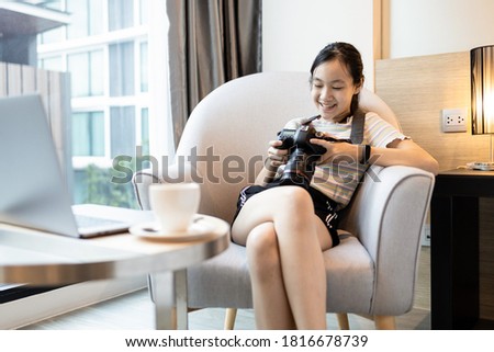 Happy smiling asian teenager girl watching photos from travel on digital camera while sitting on armchair in hotel room on summer vacations,female traveler checking picture photos after taking a photo
