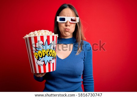Young blonde woman wearing 3d glasses and eating pack of popcorn watching a movie on cinema puffing cheeks with funny face. Mouth inflated with air, crazy expression.