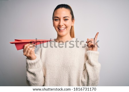 Beautiful blonde woman with blue eyes holding red paper airplane over white background surprised with an idea or question pointing finger with happy face, number one