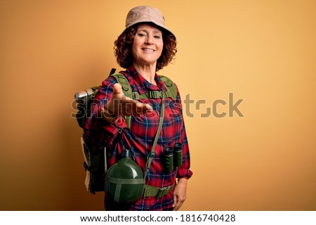 Middle age curly hair hiker woman hiking wearing backpack and water canteen using binoculars smiling cheerful offering palm hand giving assistance and acceptance. Royalty-Free Stock Photo #1816740428