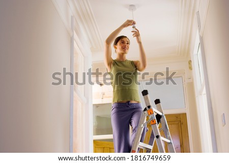 Low angle view of a woman doing DIY at home standing on ladder and attaching light bulb to the ceiling fixture.