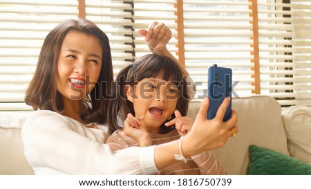 Happy Asian Family mum and little kid daughter using a smart phone and having a video call chatting or taking a selfie sitting on a couch fun and enjoying online communication in living room at home.