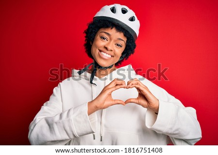 Young African American afro cyciling woman with curly hair wearing bike security helmet smiling in love showing heart symbol and shape with hands. Romantic concept.