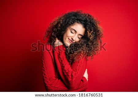 Young beautiful woman with curly hair and piercing wearing casual red sweater Hugging oneself happy and positive, smiling confident. Self love and self care