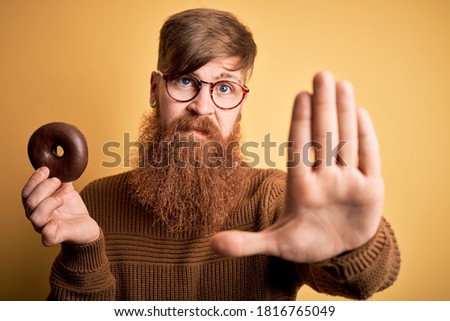 Redhead Irish man with beard eating chocolate donut over yellow background with open hand doing stop sign with serious and confident expression, defense gesture