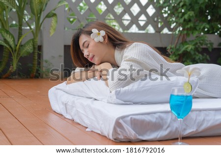 Asian Thai young woman lying and relaxing on bed near swimming pool in spa salon with a drink glass, beauty and body care concept. Outdoor spa resort. Lifestyle healthy people.