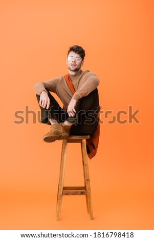 stylish man in autumn outfit and glasses sitting on wooden stool and looking at camera on orange