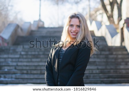 Young woman, blonde, European descent. Woman posing in a black jacket, on the stairs of concrete in a city park. Woman about 21 years. Photo taken in the cold season
