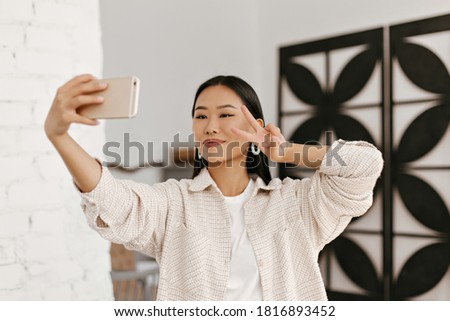 Pretty brunette woman in beige jacket and white t-shirt takes selfie and shows v-sign. Young Asian lady holds phone and smiles in cozy room.