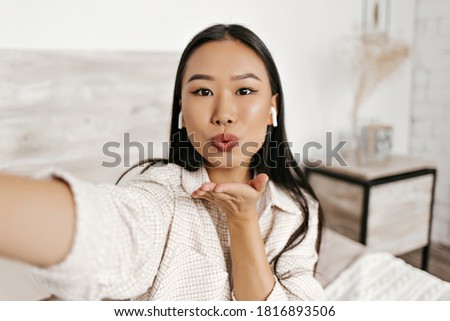 Brown-eyed brunette woman in plaid jacket takes selfie, blows kiss and listens to music in wireless headphones in cozy bedroom.