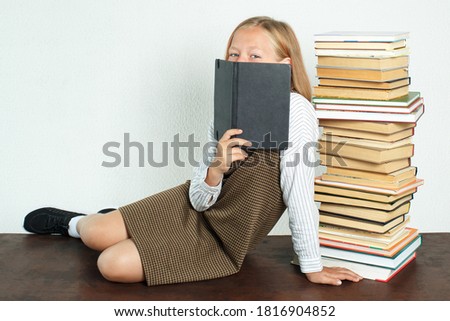 Education concept. A teenage girl sits on a table near books, covering her face with a notebook.