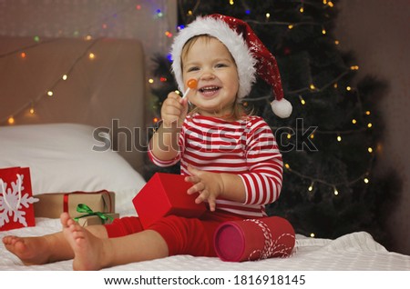 Happy Holidays! Adorable baby girl under Christmas tree with gift boxes. Christmas present. Cure girl in Santa hat open gift box.