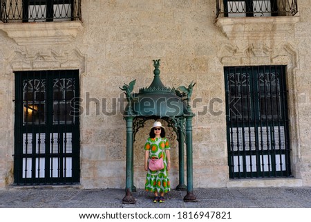 An Asian female tourist standing under an ornated broze pavilion in front of a stone building in old town Havana.