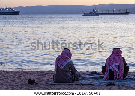 Two Arabic men wearing keffiyeh, agal and thobe are seen on the beach by the Gulf of Aqaba, sitting cross legged on a rug. They look at the beautiful sunset over red sea.