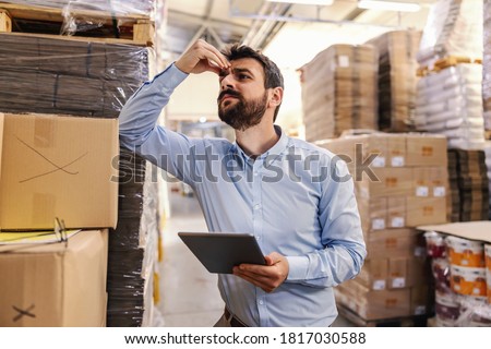 Nervous young attractive bearded businessman leaning on box in warehouse, holding tablet and thinking about problem while holding his head.