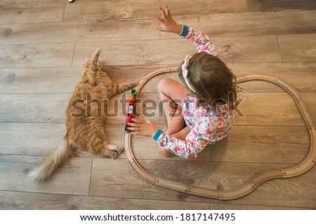 Little kid girl playing with pet cat while sitting on floor at home. Child playing toy wooden train together with ginger cat. Lifestyle child photo