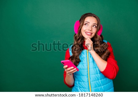 Photo of charming cute young lady look empty space finger chin think news choosing best shopping option wear pink ear warmers blue zipped vest red pullover isolated green color background