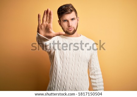 Young blond man with beard and blue eyes wearing white sweater over yellow background doing stop sing with palm of the hand. Warning expression with negative and serious gesture on the face.