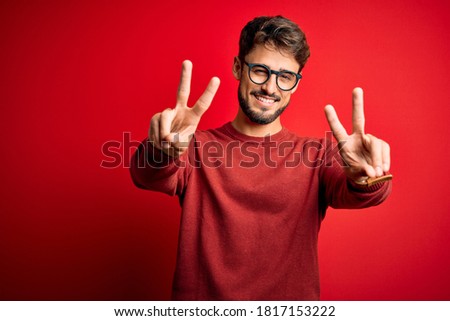 Young handsome man with beard wearing glasses and sweater standing over red background smiling with tongue out showing fingers of both hands doing victory sign. Number two. Royalty-Free Stock Photo #1817153222