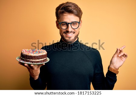 Young man with beard wearing glasses holding birthday cake over isolated yellow background very happy pointing with hand and finger to the side