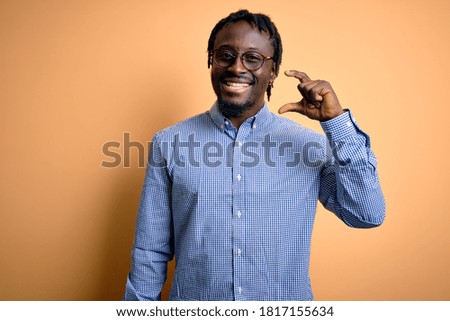 Young handsome african american man wearing shirt and glasses over yellow background smiling and confident gesturing with hand doing small size sign with fingers looking and the camera. Measure.