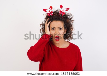 Embarrassed Young arab woman with curly hair wearing christmas headband standing on white background, with shocked expression, expresses great amazement, Puzzled girl poses indoor