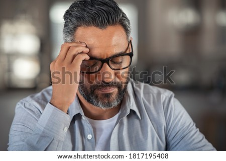 Frustrated middle aged man sitting on couch at home. Close up face of stressed indian businessman wearing eyeglasses with eyes closed. Overworked middle eastern business man with terrible migraine. Royalty-Free Stock Photo #1817195408