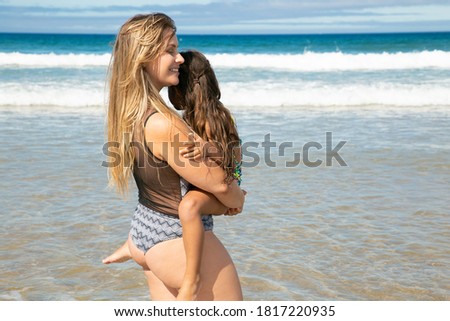Happy relaxed young mom holding little daughter in arms, carrying kid, standing on beach. Bright ocean with waves and white foam, clear sky in background. Parenting and outdoor activities concept