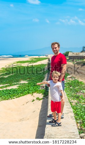 Father son walk along narrow concrete path, sea blue clear sky green grass background. Symbol as dad leads child into future adulthood. Happy childhood, fathers day, daddy influence on boy worldview