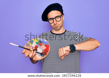 Young artist man with blue eyes wearing french beret and glasses drawing using paintbrush with angry face, negative sign showing dislike with thumbs down, rejection concept