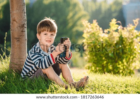 Cheerful child sitting on the grass looks cartoons in the phone in the summer at sunset. Cute boy having fun in nature