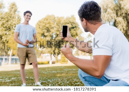 selective focus of man holding smartphone and taking photo of teenager son in park