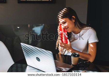 Happy young woman opening gift in front of laptop during video call or chat, celebrating birthday online. Concept of distance relations, celebrations and lifestyle.