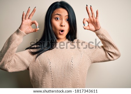 Young beautiful chinese woman wearing casual sweater over isolated white background looking surprised and shocked doing ok approval symbol with fingers. Crazy expression