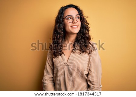 Beautiful woman with curly hair wearing striped shirt and glasses over yellow background looking away to side with smile on face, natural expression. Laughing confident. Royalty-Free Stock Photo #1817344517