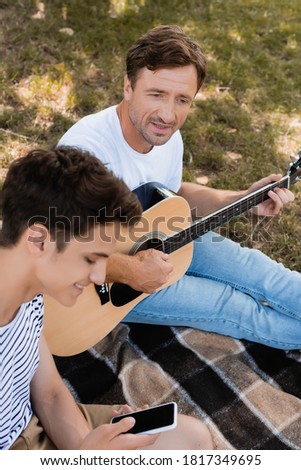 selective focus of father sitting on blanket and playing acoustic guitar near teenager son holding smartphone