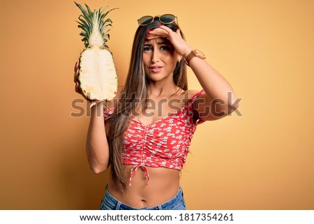 Young beautiful brunette woman holding middle pineapple fruit over isolated yellow background stressed with hand on head, shocked with shame and surprise face, angry and frustrated. Fear and upset.