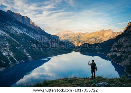 Mobile phone selfie hiker taking photo or video with cellphone in Oeschinensee Lake banner panorama background. Nature walk recreational activity man happy using tech device. Outdoor lifestyle.