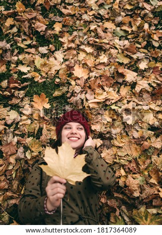 Young beautiful woman in a burgundy knitted hat and scarf holds yellow maple leaves laying in an autumn park with colored, red and yellow leaves on the trees. Stylish autumn clothes, inspiration, mood