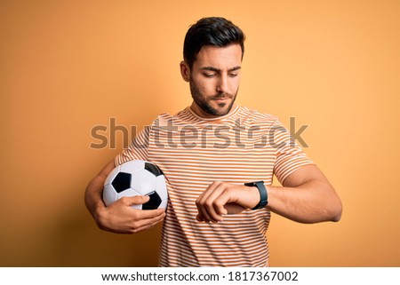 Handsome player man with beard playing soccer holding footballl ball over yellow background Checking the time on wrist watch, relaxed and confident