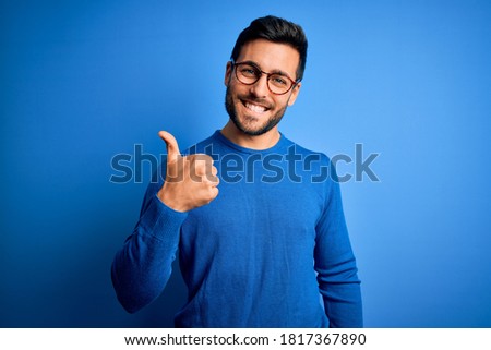 Young handsome man with beard wearing casual sweater and glasses over blue background doing happy thumbs up gesture with hand. Approving expression looking at the camera showing success. Royalty-Free Stock Photo #1817367890