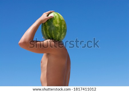 Back view of a shirtless male holding a watermelon on the shoulder with the sky in the background.
