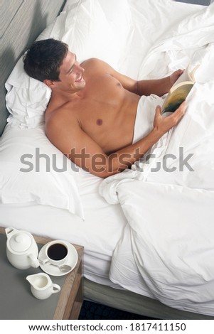 Top view of a shirtless man reading a book while lying on the bed.