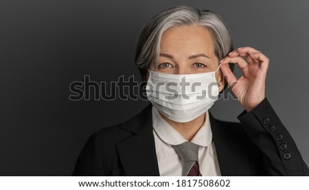 Pretty gray-haired middle-aged business woman putting on protective mask and showing ok or okey sign while posing on gray wall background. Quarantine concept.