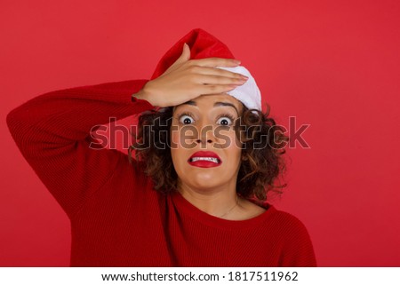Oops, what did I do? Beautiful woman wearing a Christmas hat  holding hand on forehead with frightened and regret expression. Wearing casual clothes standing outdoors.