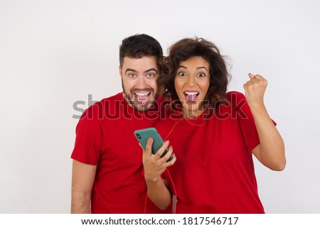 Positive Young beautiful couple wearing red t-shirt on white background holds modern cell phone connected to headphones, clenches fist from good emotions, exclaims with joy, 