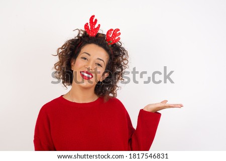 Young arab woman with curly hair wearing christmas headband standing on white background smiling cheerful presenting and pointing with palm of hand looking at the camera.